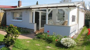 Uma casa pequena, térrea, com varanda coberta, um pequeno jardim com flores e uma chaminé visível.