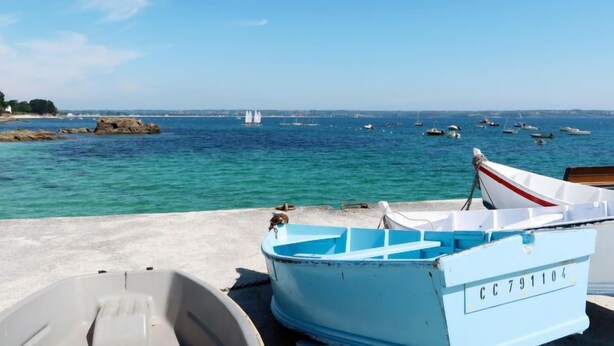 Escena costera con barcos amarrados a un muelle, cielo azul despejado y mar en calma.