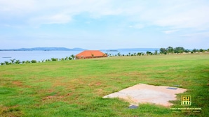 A grassy field with a building in the distance, a clear sky, and a body of water. - Fancy Resort Beach Hotel (Jinja)