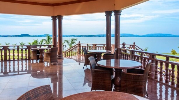 A covered outdoor area with tables and chairs, a railing, and a view of a body of water and distant land. - Fancy Resort Beach Hotel (Jinja)