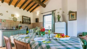 Un coin repas avec une nappe à carreaux, des chaises en bois, un saladier de fruits et un réfrigérateur en arrière-plan.