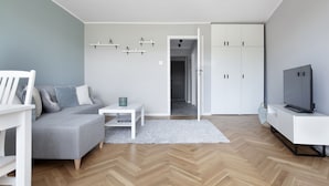 A modern living room with a grey sofa, white coffee table, and a TV on a white cabinet. There is a hallway leading to a wardrobe with doors. - Elite Apartments Przymorze (Gdansk)