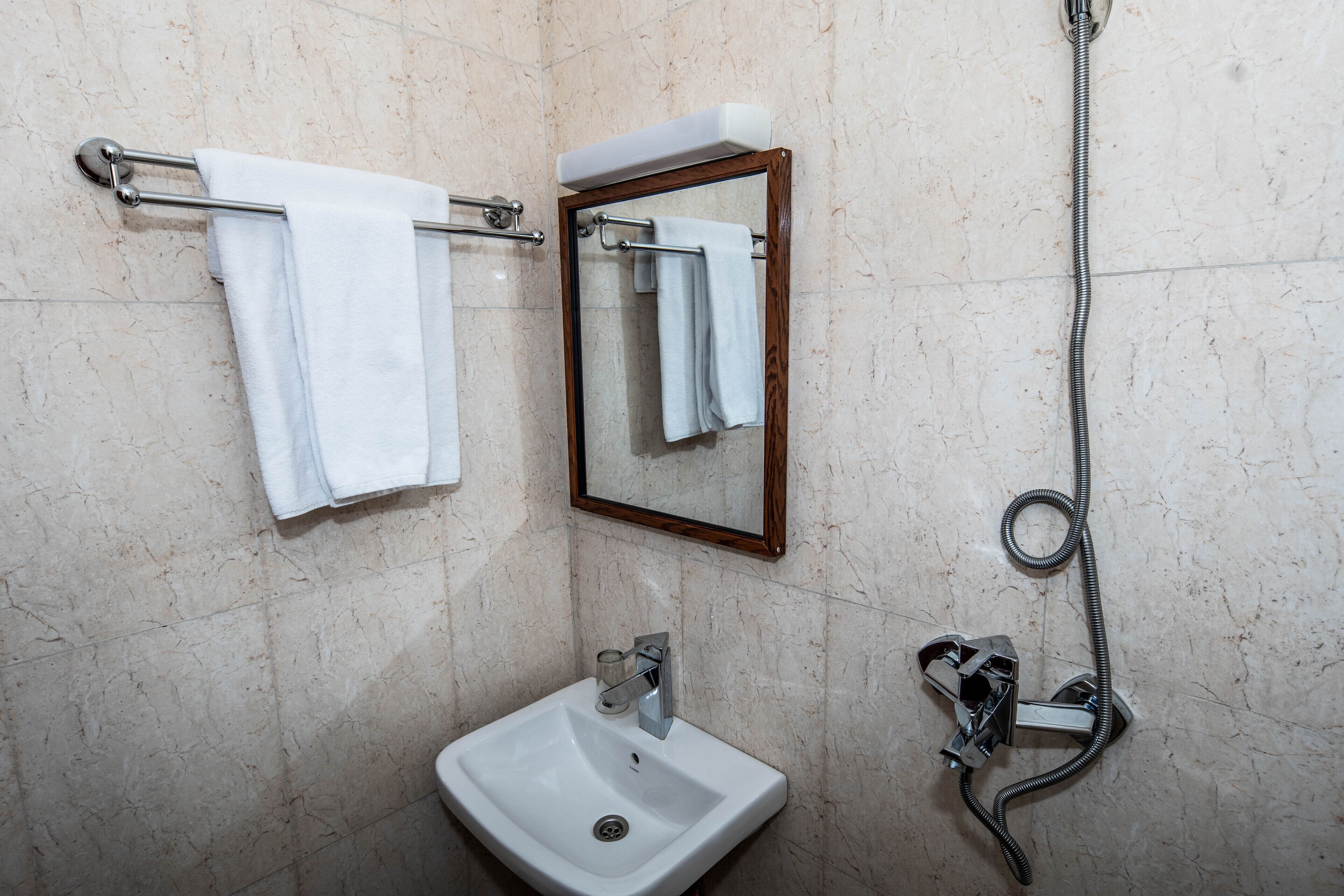 A bathroom with a white sink, a towel rack with two towels, a mirror with a wooden frame, and a shower hose.