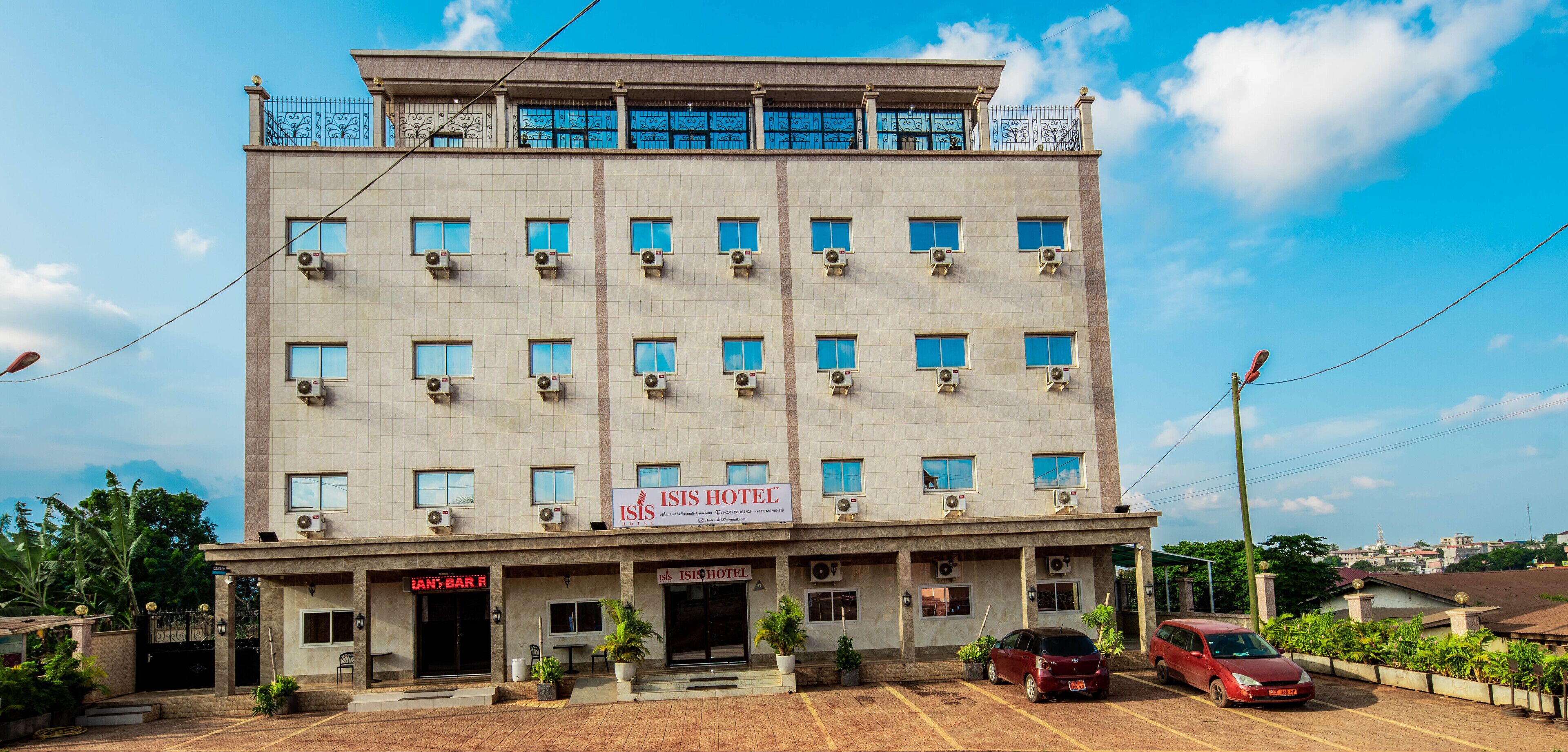 A multi-story hotel with a sign for "Isis Hotel" and "Ian's Bar R." There are air conditioning units on the facade, a parking lot with cars, and a clear blue sky in the background.