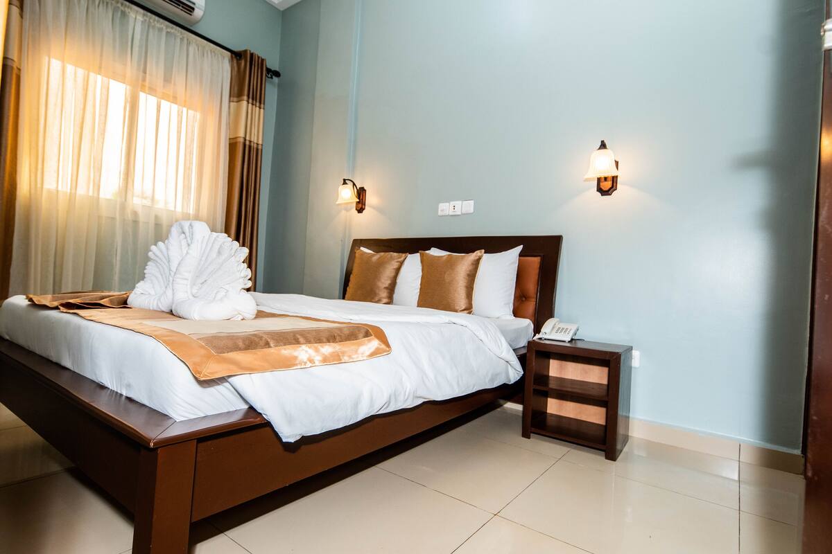 A neatly made bed with a brown headboard, a bedside table with a lamp, and a window with sheer curtains.
