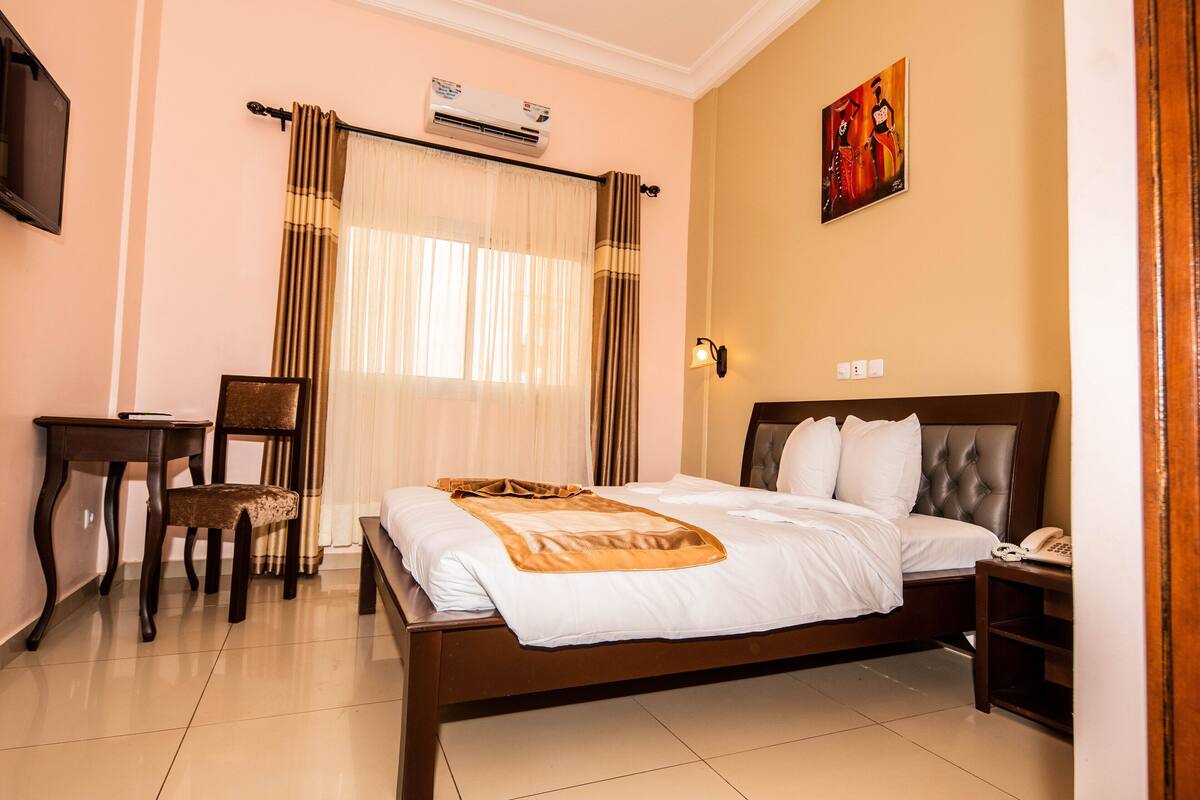 A hotel room with a bed, a desk, a chair, a wall-mounted phone, and an air conditioning unit.
