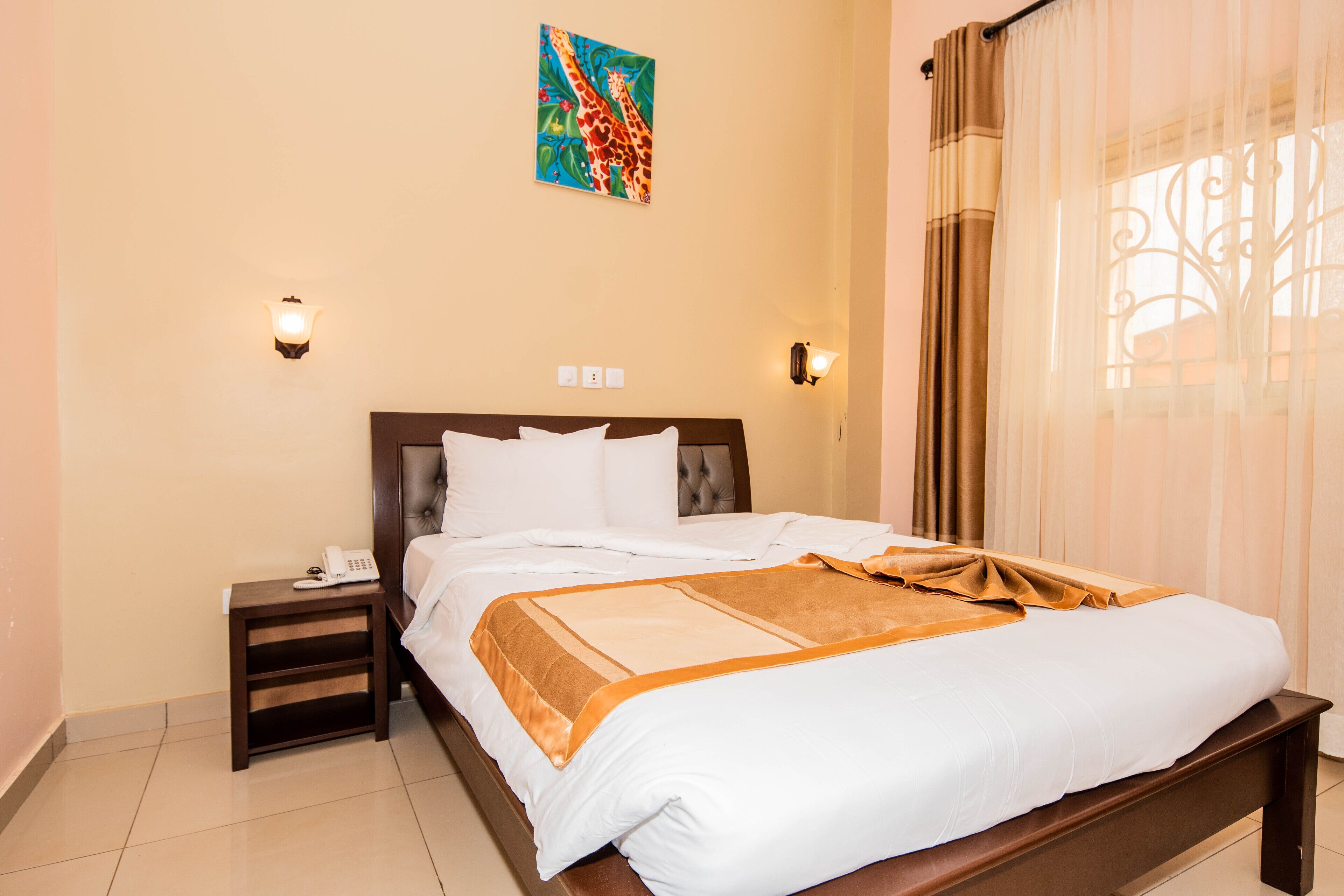 Quarto de hotel com uma cama grande, mesa de cabeceira com telefone, duas luminárias na parede e uma janela com cortinas.