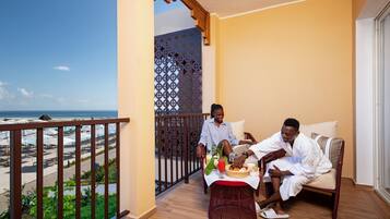 A balcony with a view of the beach, a table with food and drinks, and two people sitting on a couch.