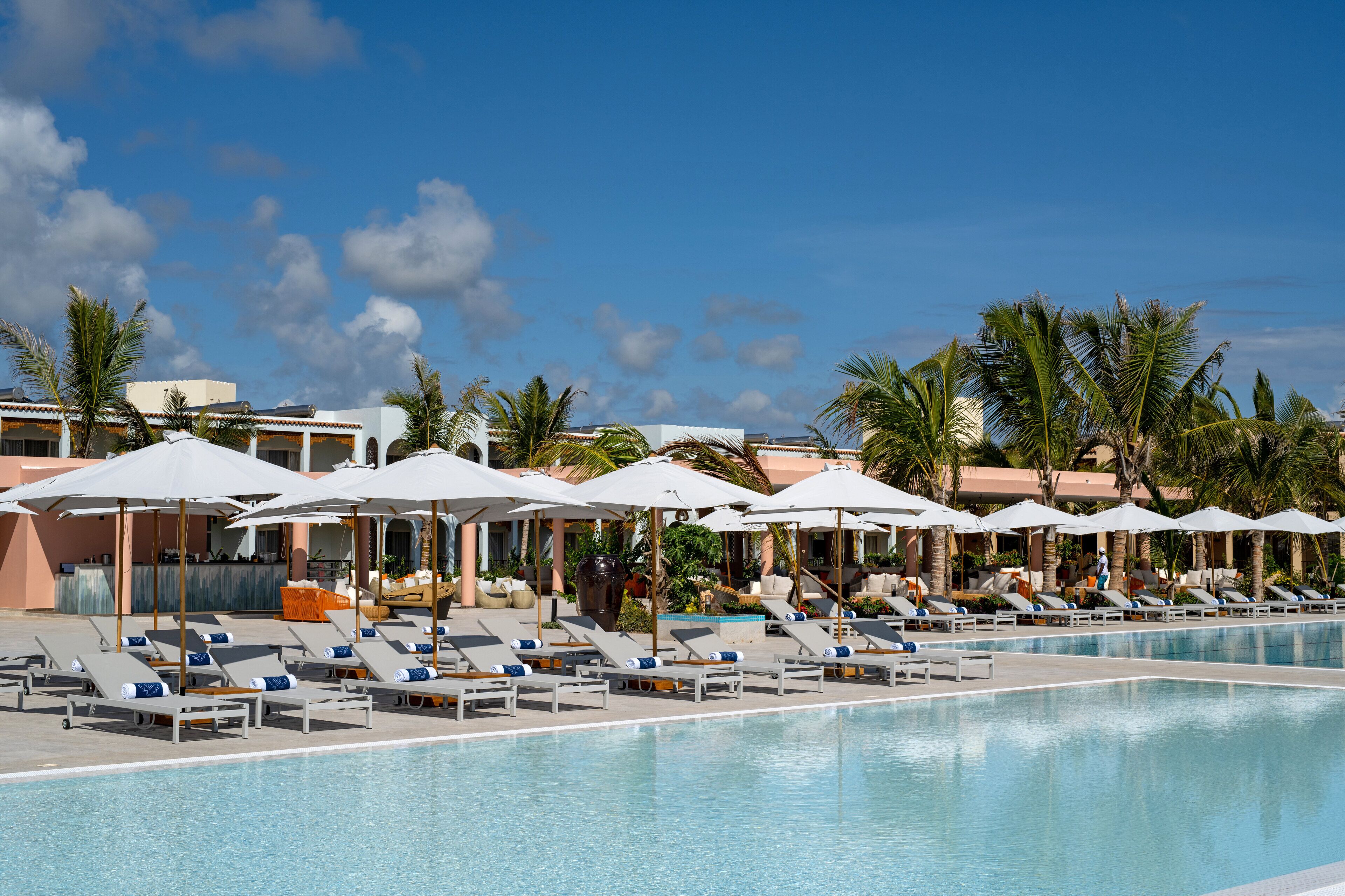 A pool area with lounge chairs and umbrellas, surrounded by palm trees and a clear sky.