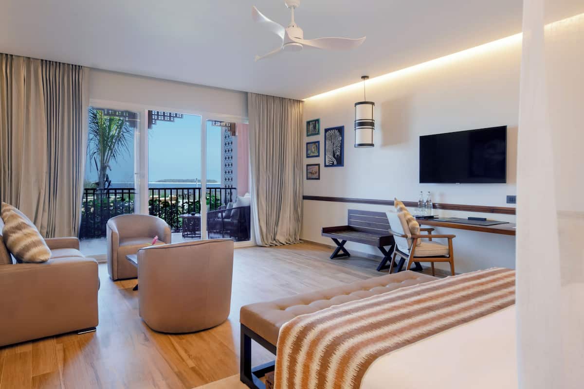 Habitación de hotel con cama, dos sillones, escritorio, televisor y balcón con vistas.