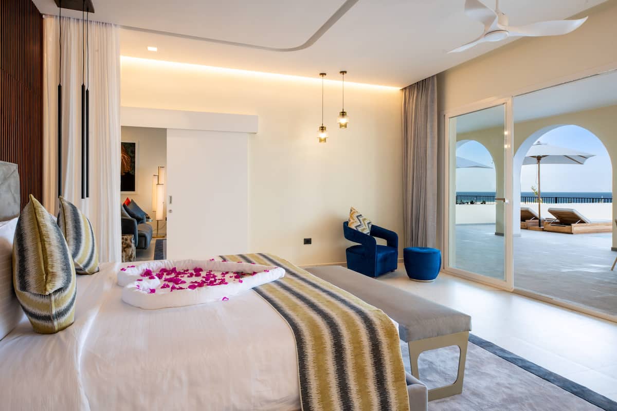 Habitación de hotel moderna con una cama grande, un banco y vista al mar a través de una puerta de cristal.