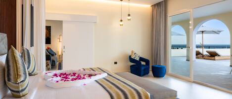 A modern hotel room with a large bed, a bench, and a view of the sea through a glass door.