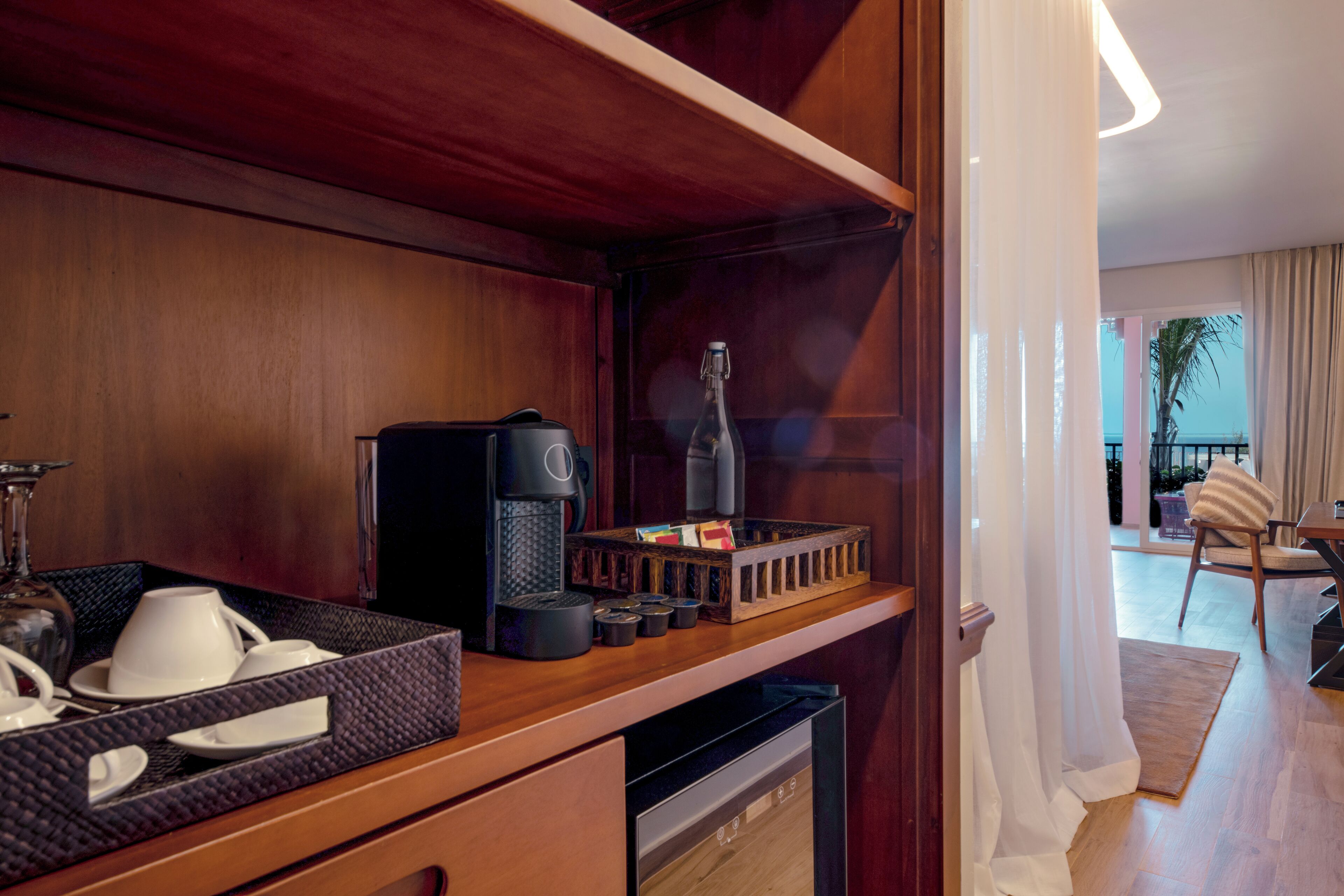 Habitación de hotel con minibar de madera, cafetera, una botella y una bandeja con tazas y platillos.