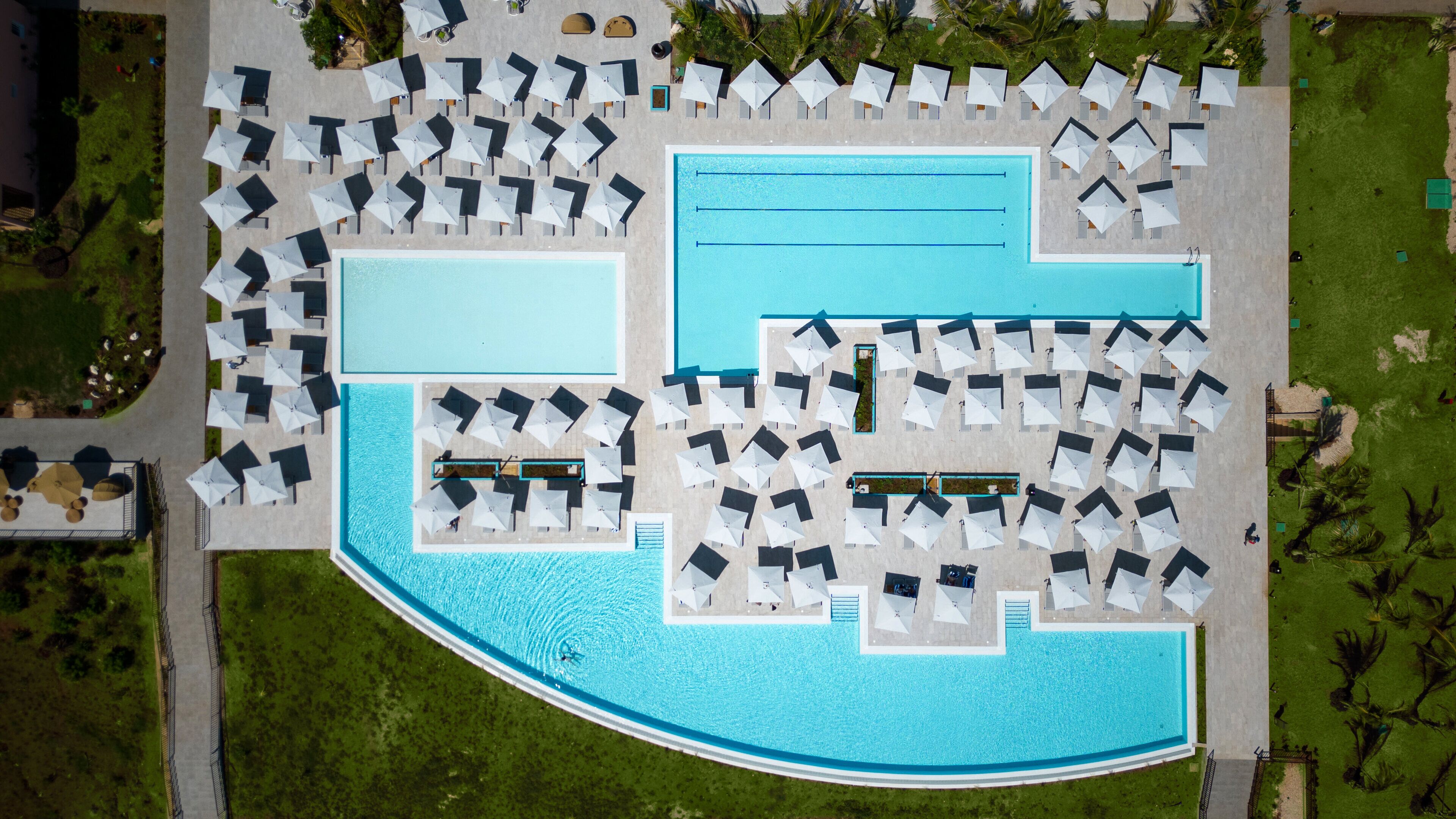 Luchtbeeld van een resort met een groot zwembad, meerdere loungegedeeltes met parasols, en een verzorgd gazon.