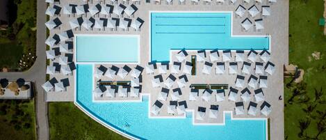 An aerial view of a resort with a large pool, multiple lounge areas with umbrellas, and a well-maintained lawn.