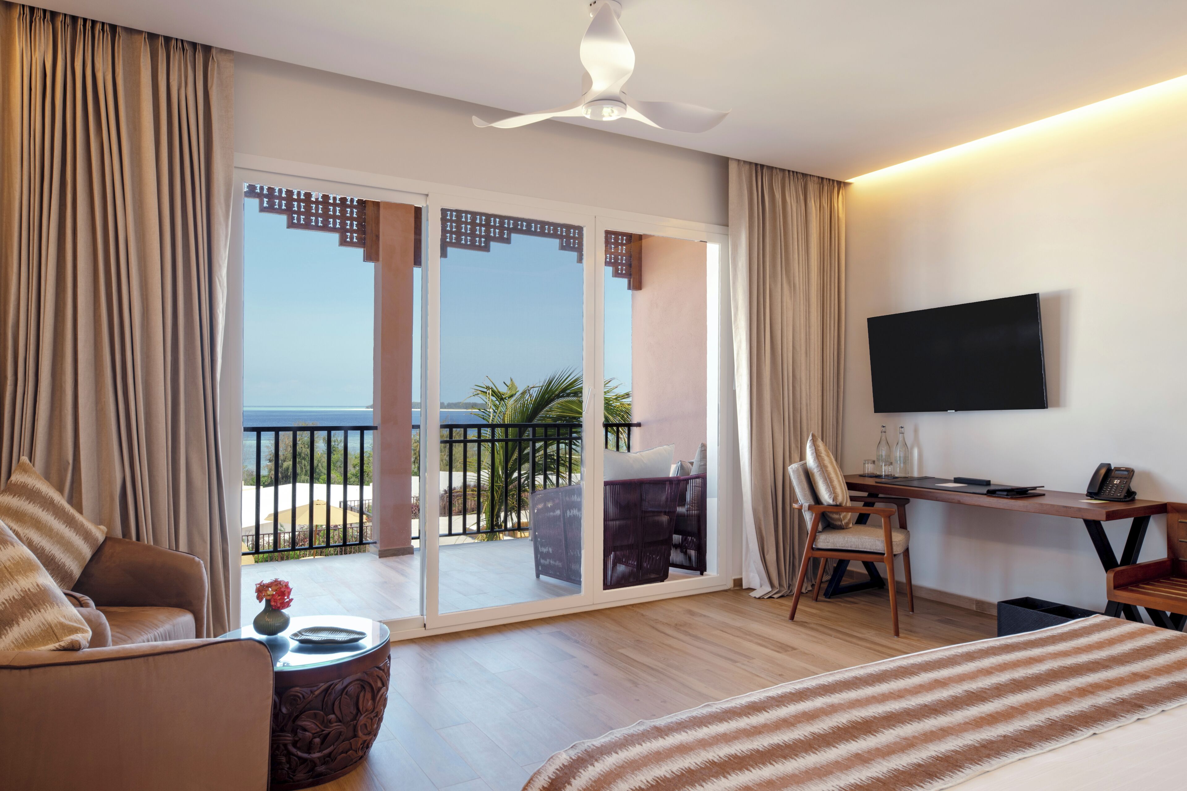 Een hotelkamer met een bed, een bureau, een stoel, een televisie en een balkon met uitzicht op zee.