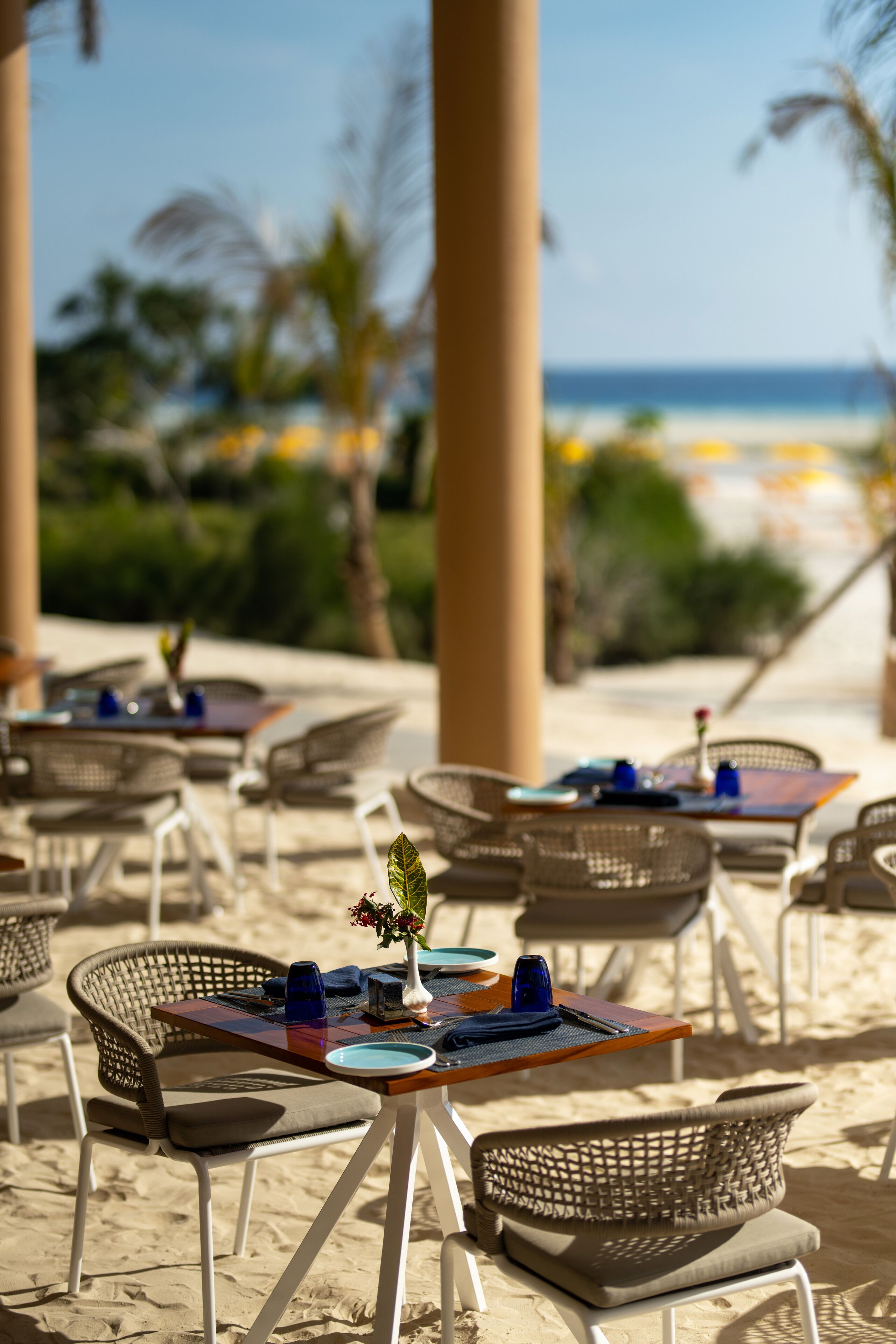 Outdoor seating with tables and chairs, a small table with a vase and a blue cup, and a view of the beach and ocean.