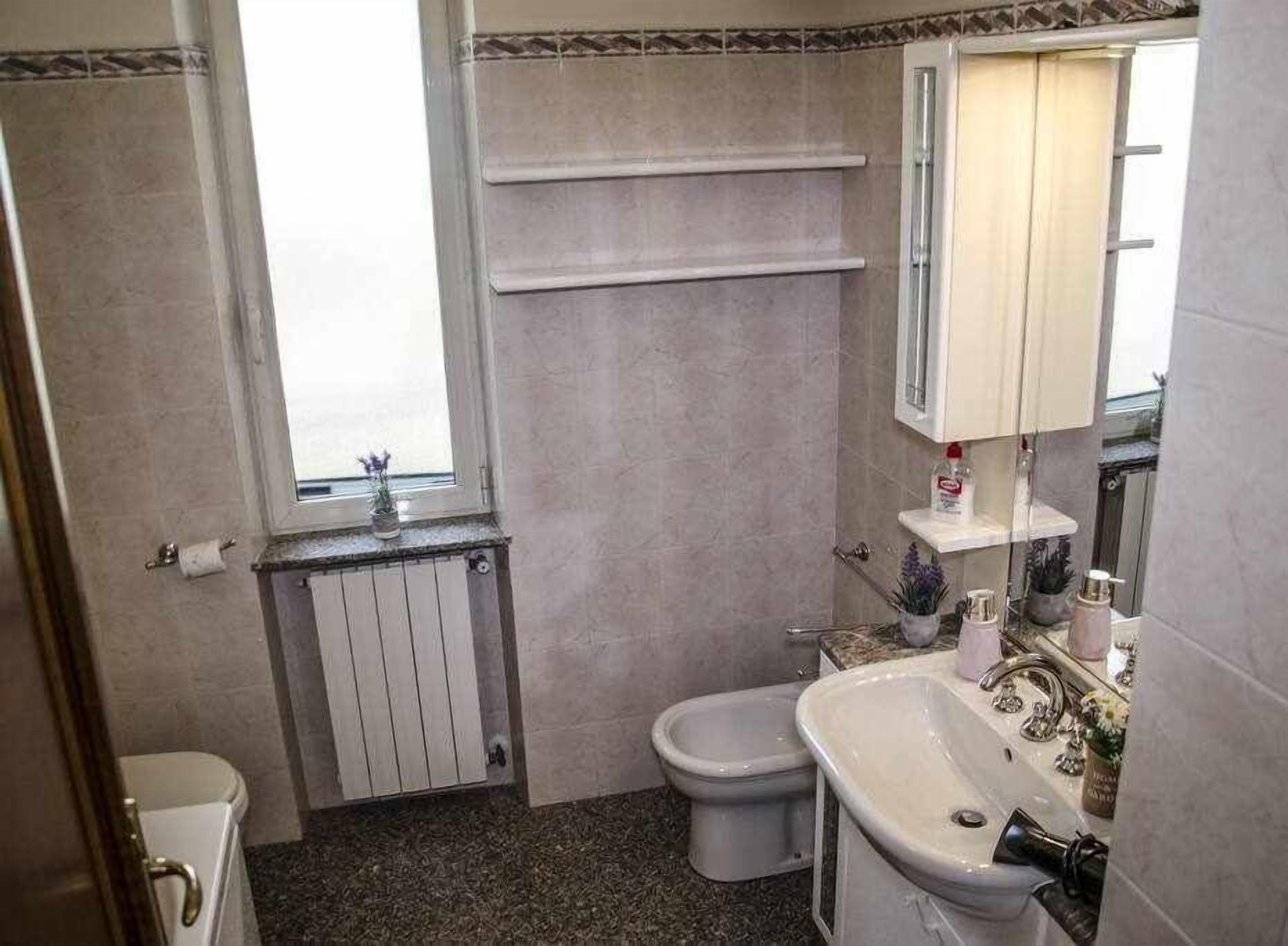 A bathroom with a toilet, bidet, and sink. There is a window with a view, a towel rack, and a shelf with toiletries.