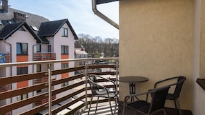 Un balcon avec des fauteuils en osier et une petite table, donnant sur une rue bordée d’immeubles et d’arbres.