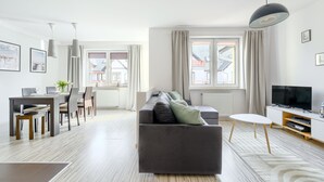 Un salón moderno con un sofá gris, una mesa de comedor con sillas, un televisor sobre un mueble bajo y ventanas amplias con cortinas.