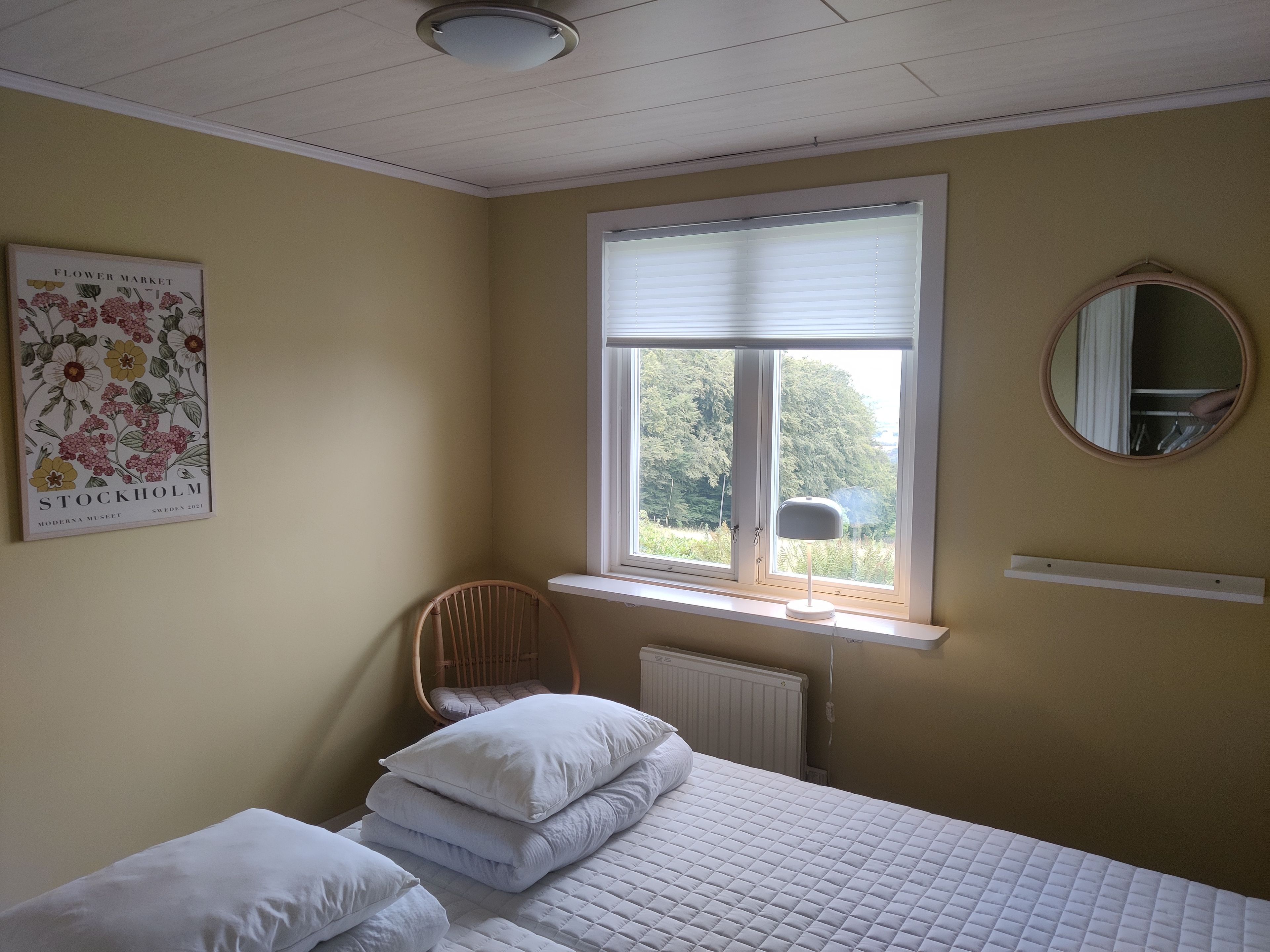Un dormitorio con cama, almohadas, una silla, ventana con persianas, radiador, espejo fijado a la pared y un cuadro enmarcado.