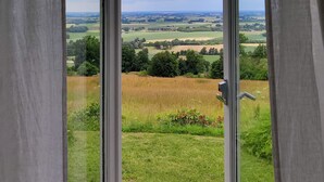 Uitzicht door een raam met gordijnen, waarop een landelijk landschap met velden en bomen te zien is.