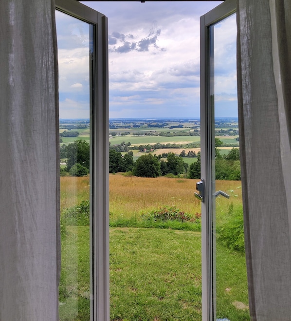 Uitzicht door een raam met gordijnen, waarop een landelijk landschap met velden en bomen te zien is.