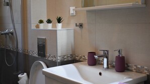 A modern bathroom with a white sink, two purple soap dispensers, a glass shower enclosure, and a small potted plant on the vanity. - Apartment Vapor (Dubrovnik)