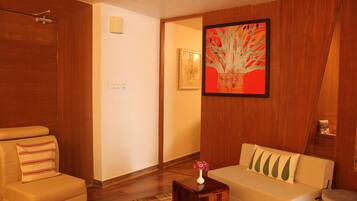 A room with a wooden floor, a beige sofa with decorative pillows, a small wooden side table with a vase, and a framed artwork on the wall.