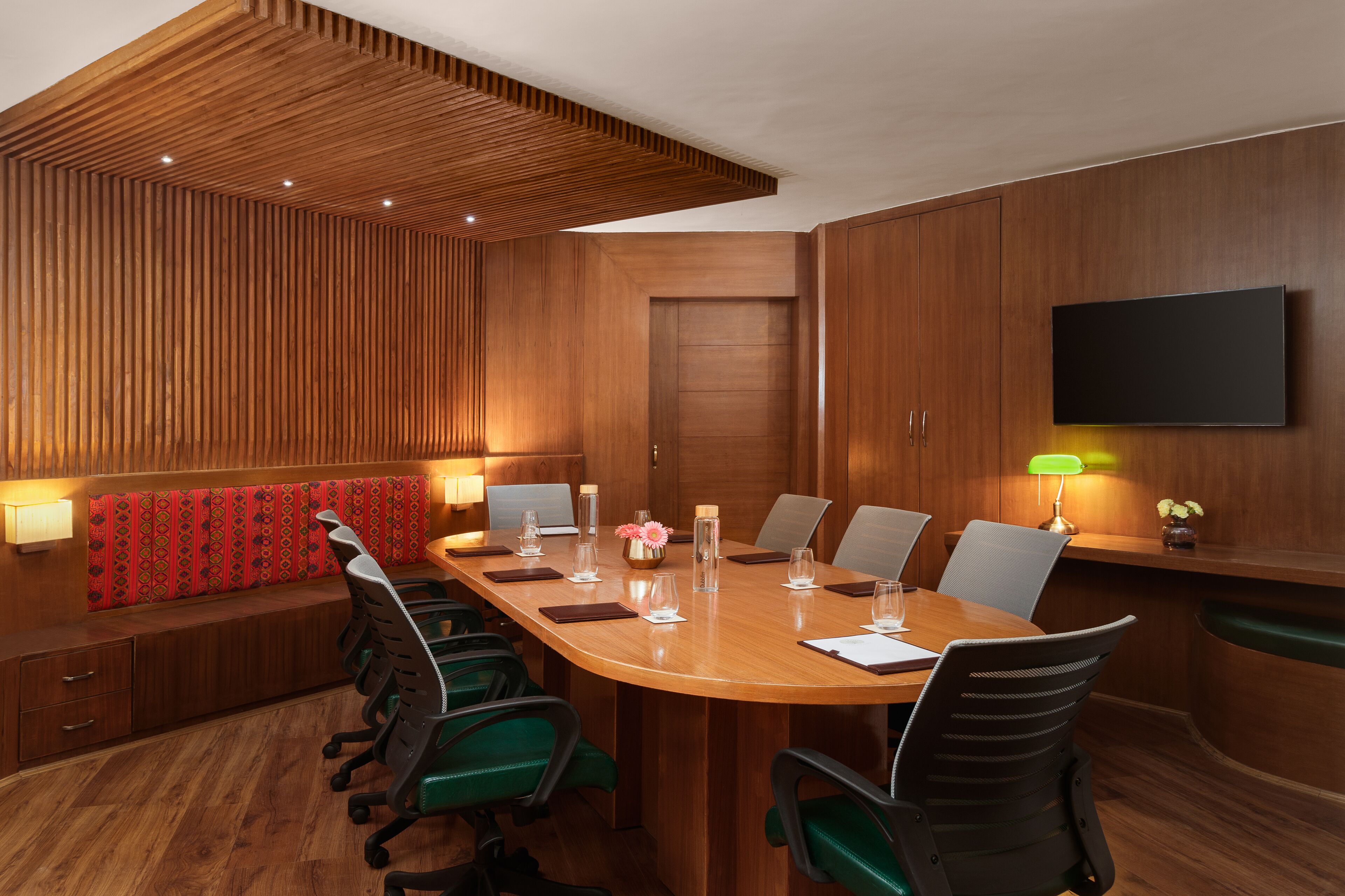 Uma sala de conferências com uma mesa de madeira, cadeiras verdes, um banco estofado vermelho e uma televisão fixada na parede.