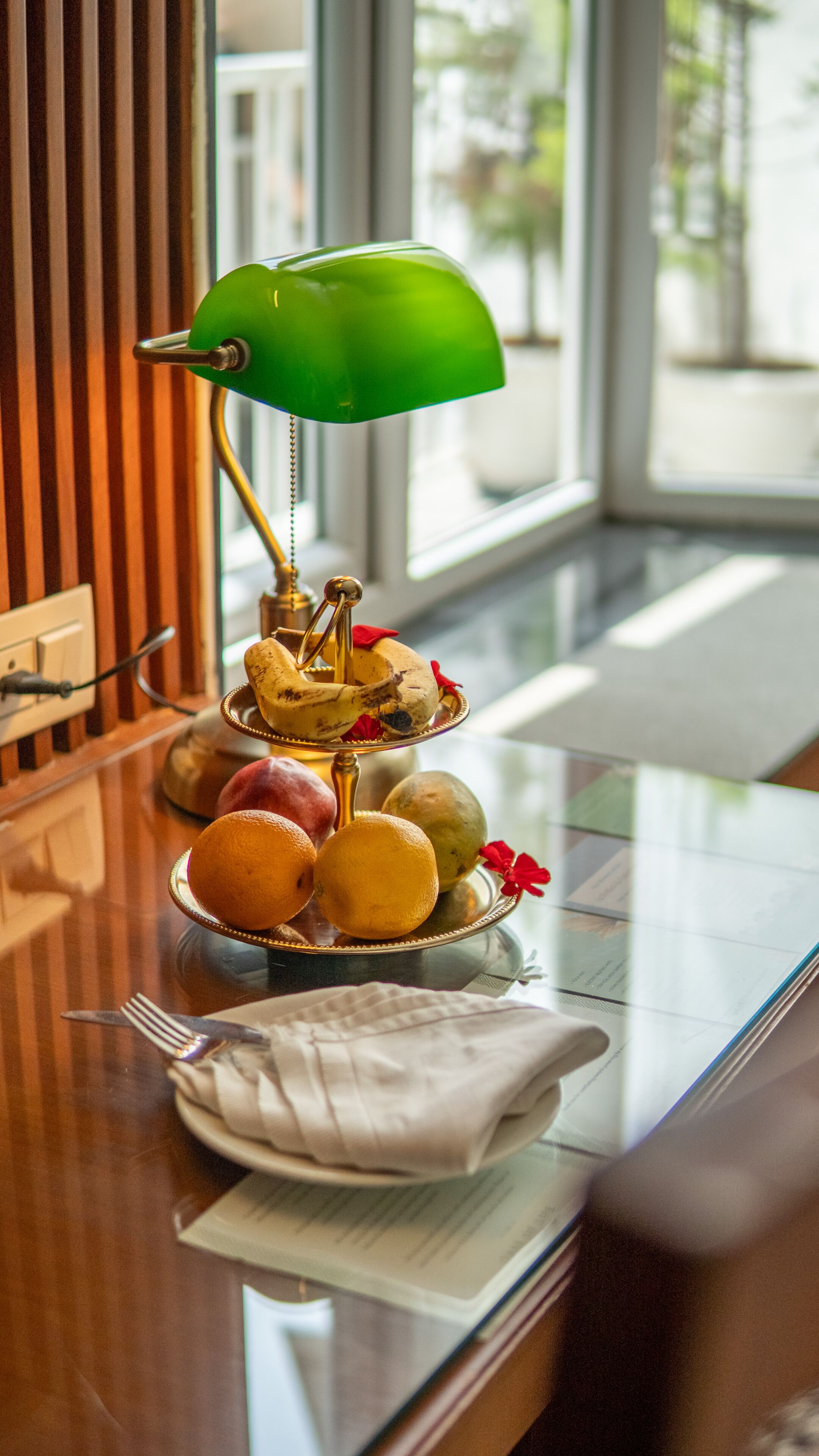 A table with a green desk lamp, a fruit tray, and a napkin on a wooden surface.
