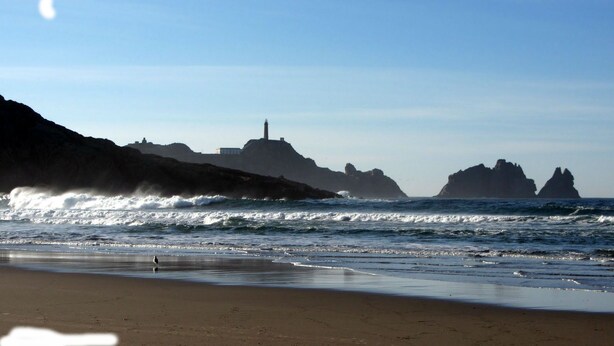 Una playa con olas, un faro y formaciones rocosas al fondo.
