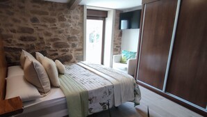 Un dormitorio con pared de piedra, una cama con almohadillas color beige, una colcha estampada con motivos florales y un armario de madera.