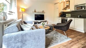 A modern living room with a grey sofa, a coffee table, and a TV. Adjacent is a kitchen with white cabinets, a microwave, and a stove. - Vacation village house 63b (Ostseebad Boltenhagen)