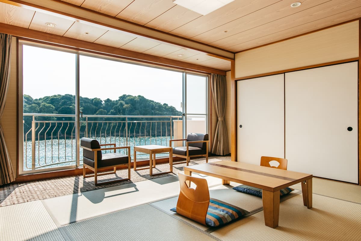 Ein Zimmer mit Tatami-Böden, einem Holztisch und Stühlen, große Fenster mit Blick auf eine bewaldete Landschaft.