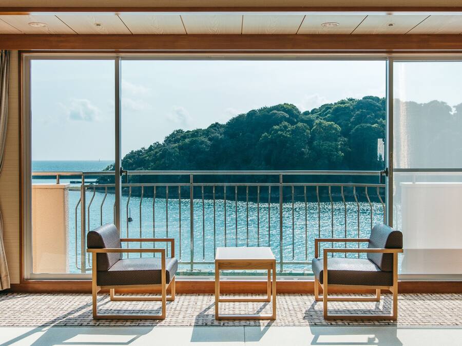 A balcony with two chairs and a small table, overlooking a body of water and a forested hill.
