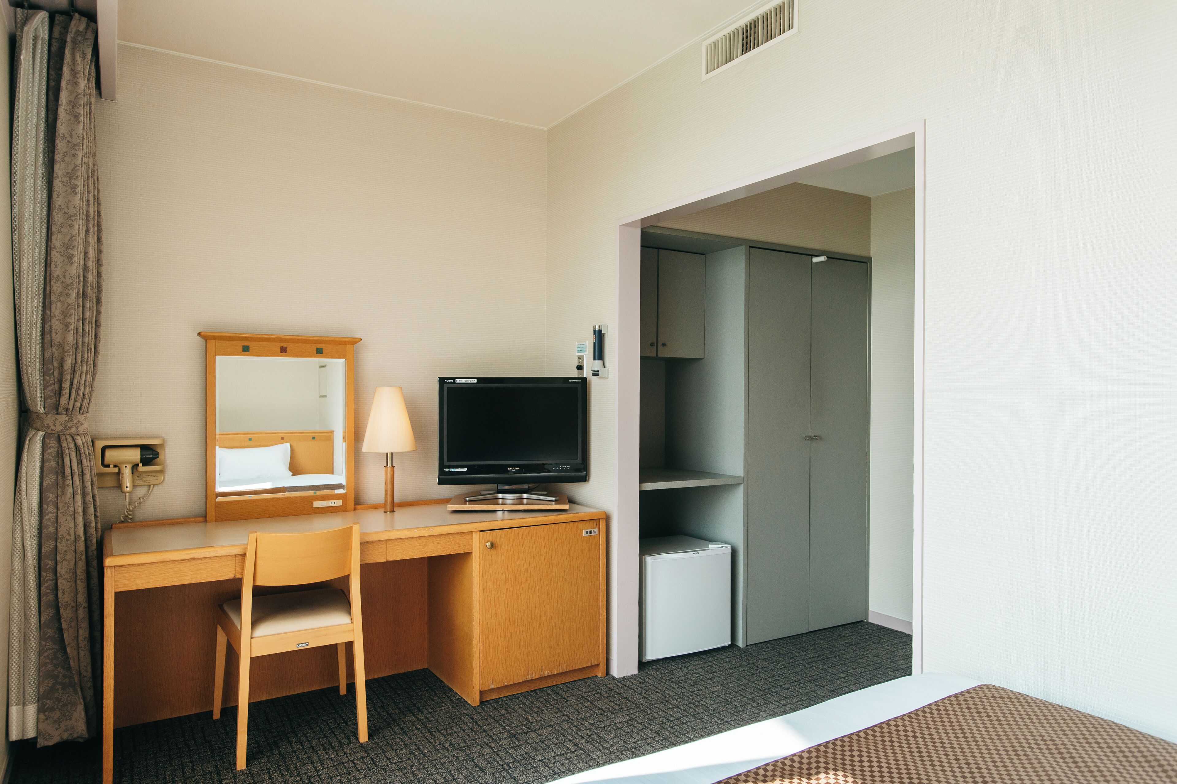 Une chambre d’hôtel équipée d’un bureau en bois, d’une télévision, d’un miroir, d’une lampe, d’une armoire et d’un petit réfrigérateur.