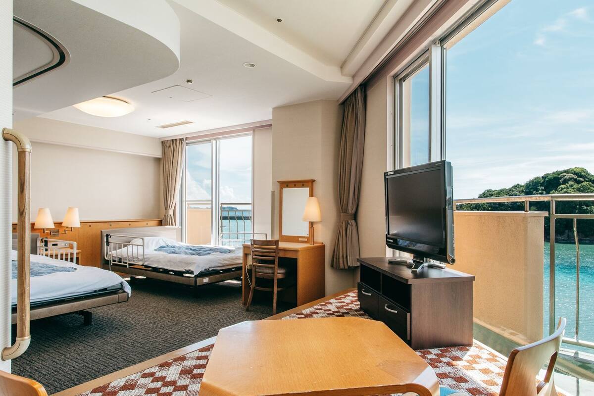 Ein Hotelzimmer mit zwei Betten, einem Fernseher, einem Schreibtisch und einem Balkon mit Meerblick.