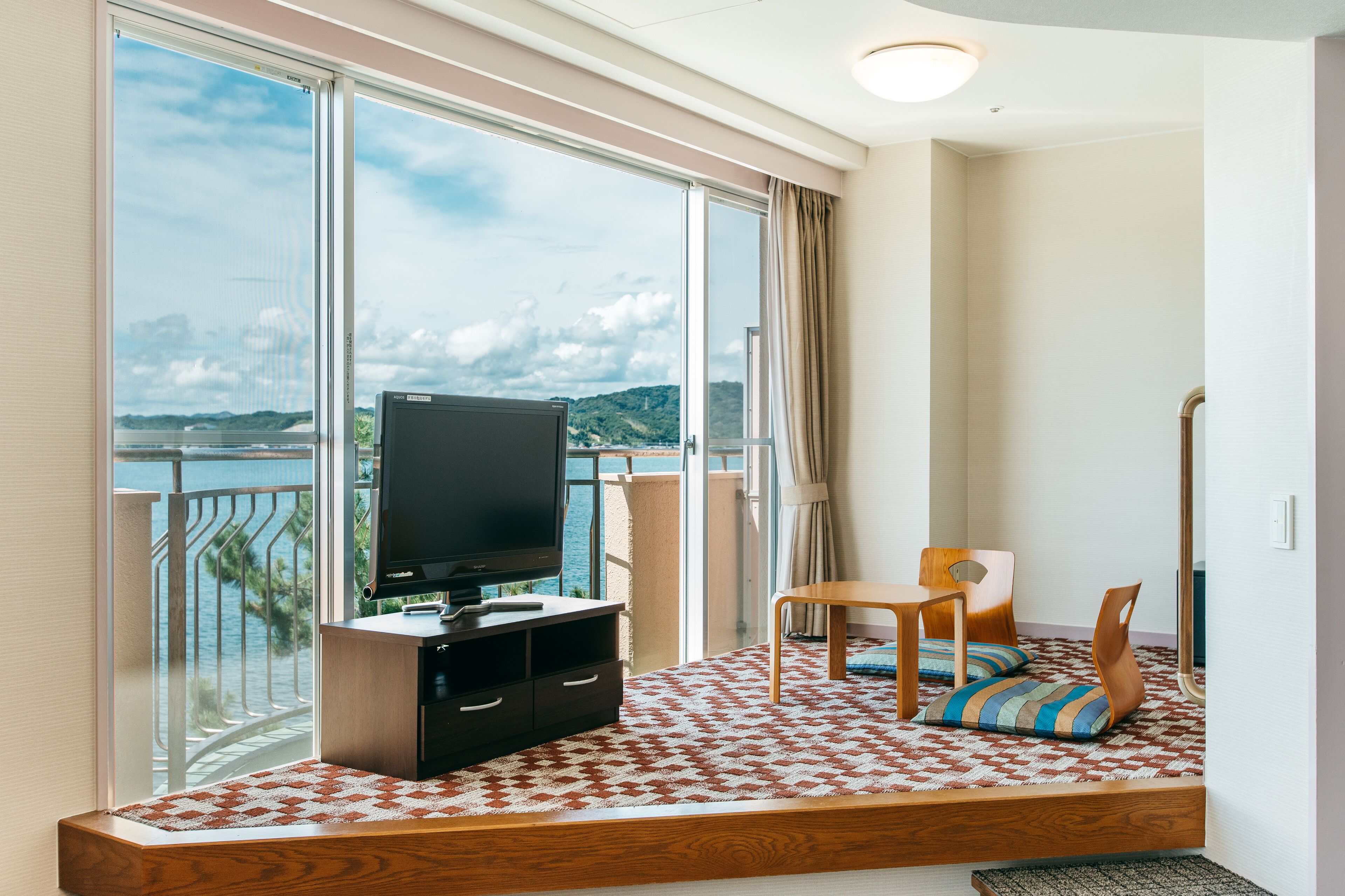 A balcony with a TV, a chair, a sofa with patterned cushions, and a view of the sea.