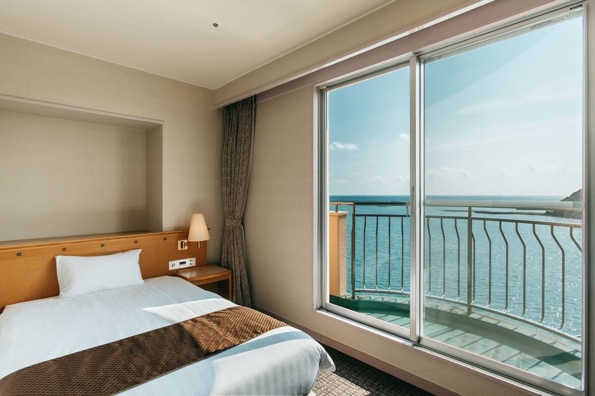 Ein Hotelzimmer mit großem Fenster, einem Bett, einem Nachttisch und Blick aufs Meer.