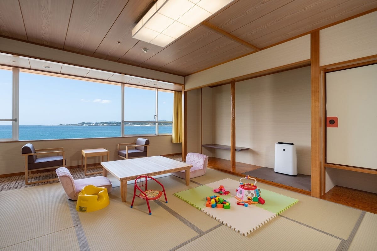 Ein Zimmer mit Tatami-Böden, einem Holztisch, einer Couch, einem Sessel und Meerblick.