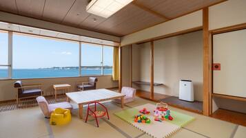 A room with tatami flooring, a wooden table, a sofa, a chair, and a view of the sea.