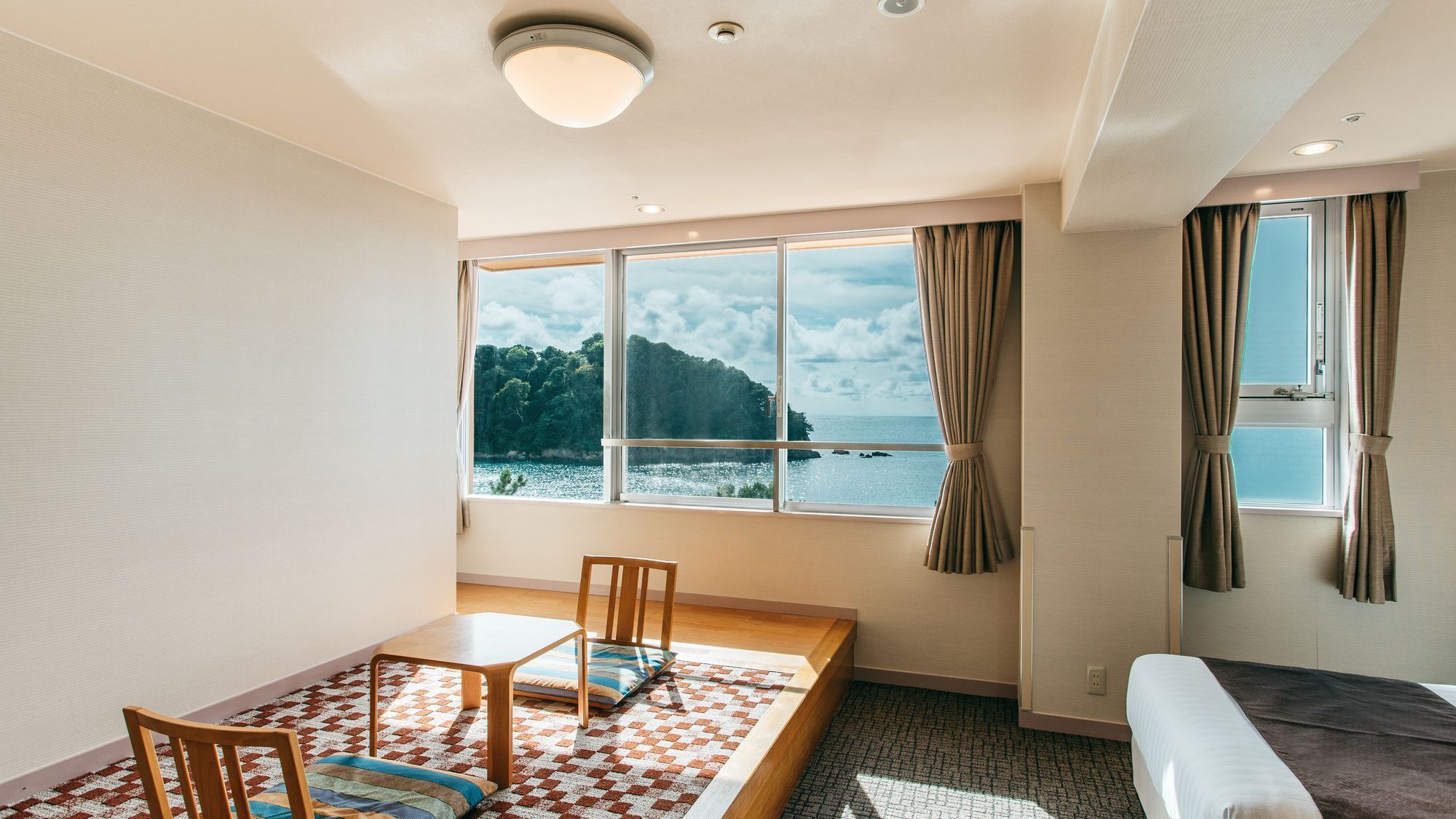 Une pièce dotée d’une grande fenêtre donnant sur la mer et une zone boisée, avec un lit, une petite table et des chaises, ainsi qu’un canapé.