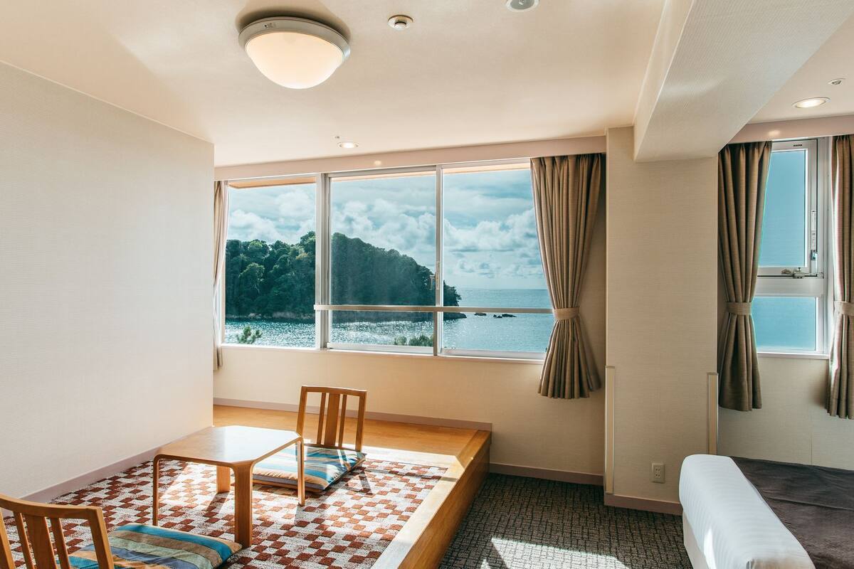 Ein Zimmer mit großem Fenster und Meer- sowie Waldblick, einem Bett, einem kleinen Tisch mit Stühlen und einem Sofa.