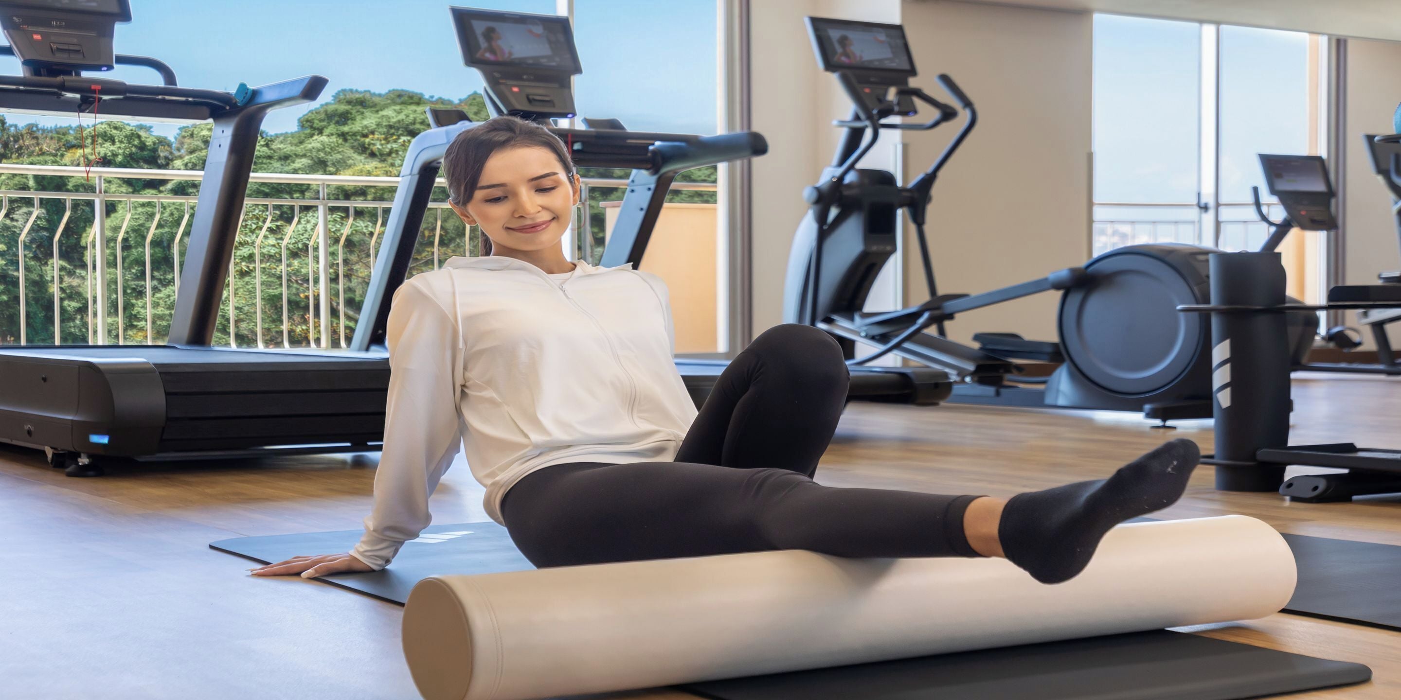 A gym with exercise equipment, a person sitting on a mat, and a large window with a view of trees.
