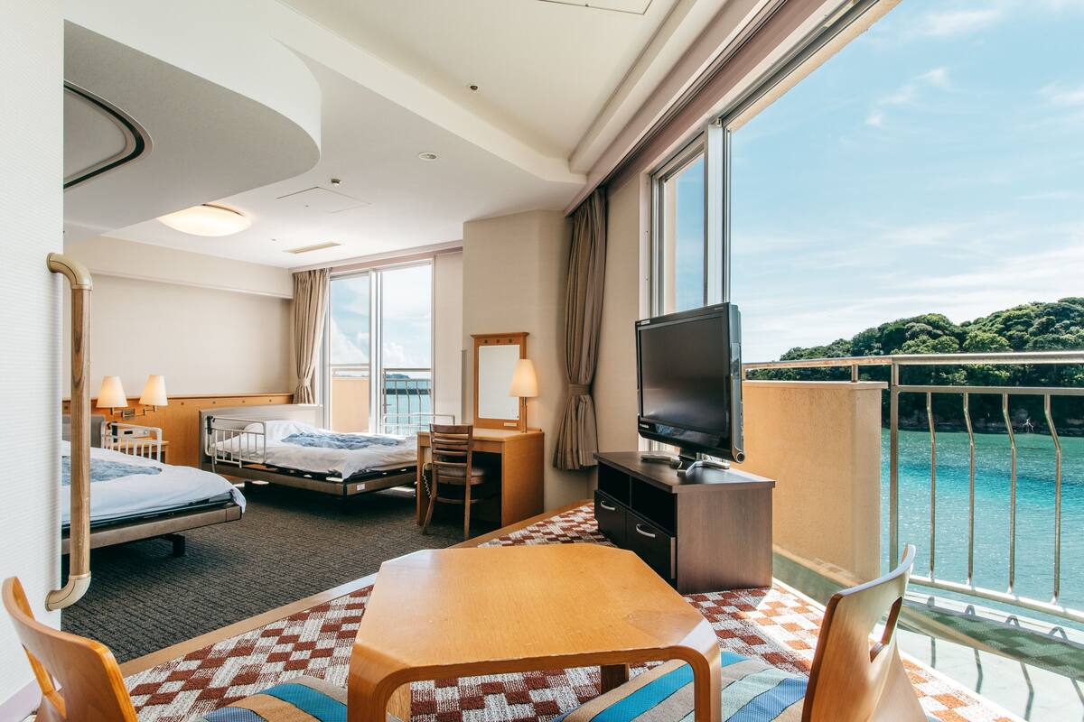 Ein Hotelzimmer mit zwei Betten, einem Fernseher, einem Schreibtisch und einem Balkon mit Blick auf das Wasser.