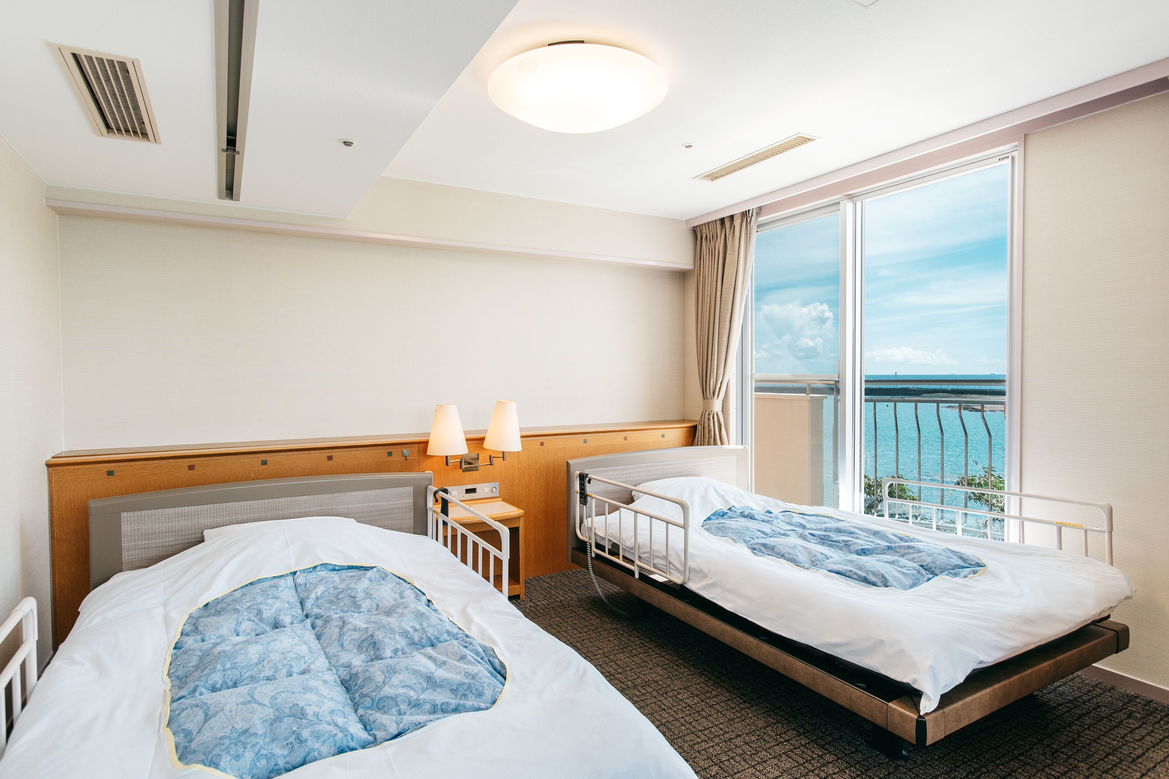 A hotel room with two beds, a window with a view of the sea, a bedside table with lamps, and a ceiling light.