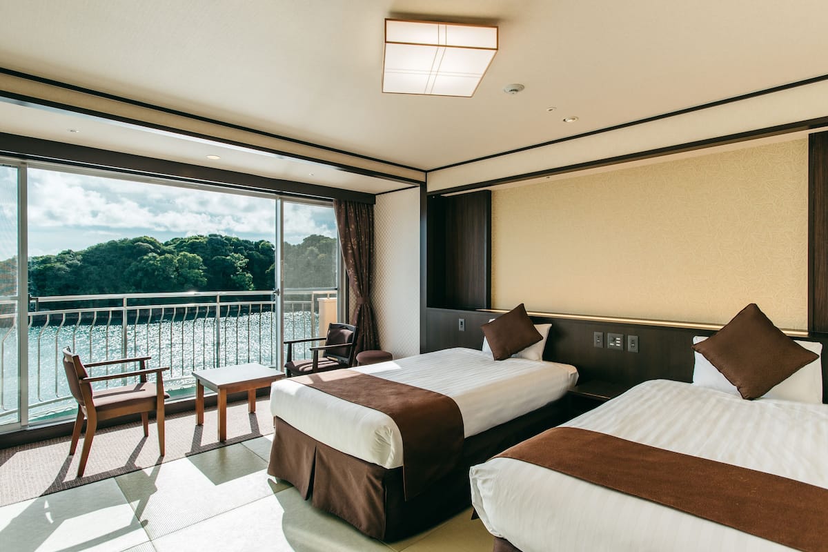Ein Hotelzimmer mit zwei Betten, einem großen Fenster mit Blick auf ein Gewässer, einem Tisch und Stühlen.
