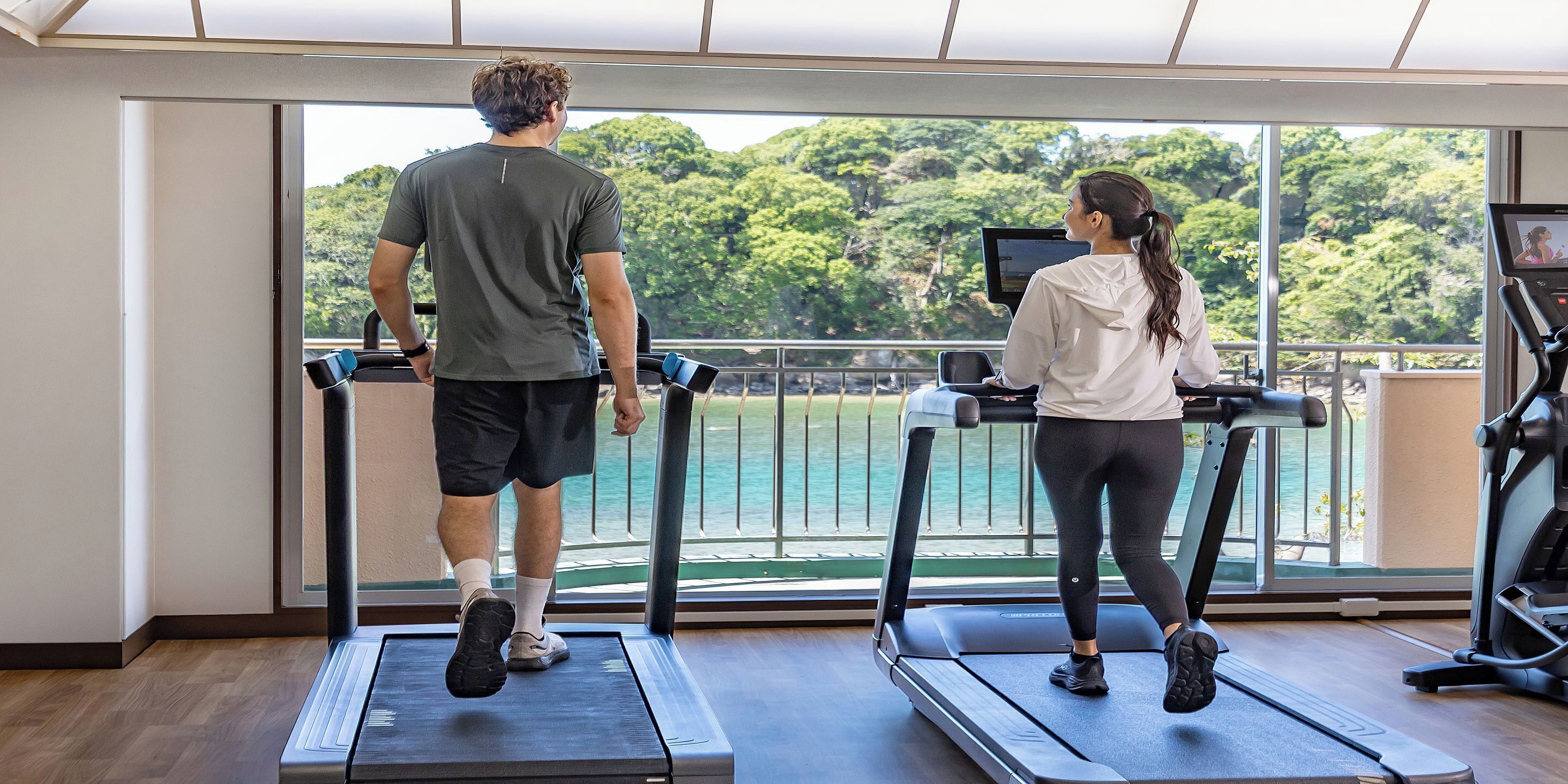 Two people on treadmills facing a large window with a view of a body of water and trees.