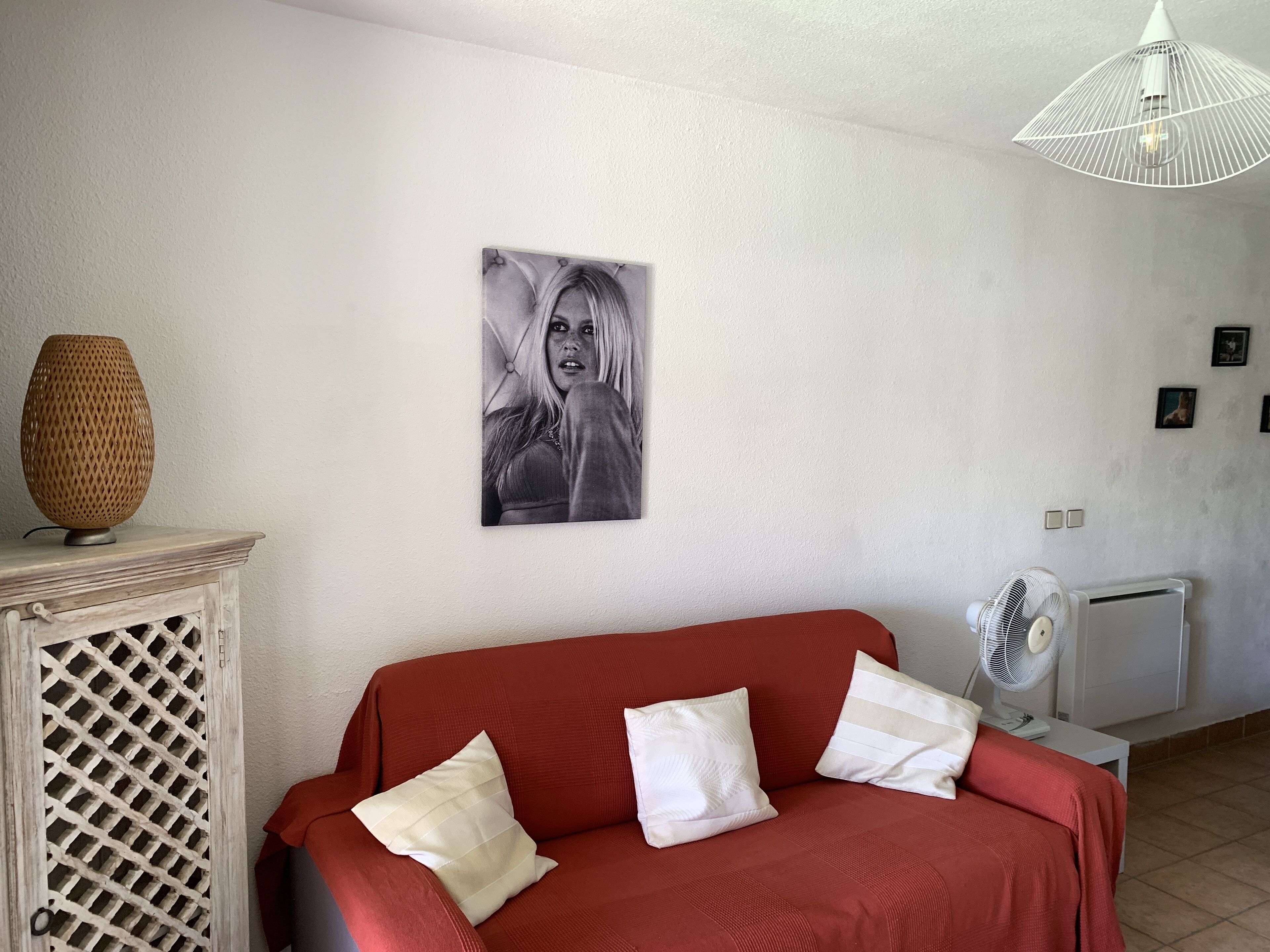 A living room with a red sofa, white cushions, a wall-mounted picture, a wicker lamp, and a white ceiling fan.